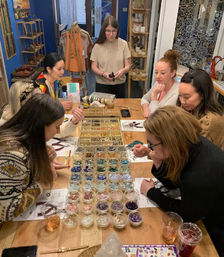 Several women at a cozy craft studio gathered around a wooden table filled with dozens of small bowls of colorful beads, gemstones, tools, and drinks during a jewelry-making workshop.