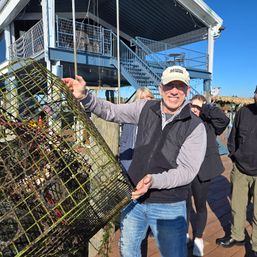 Smiling man lifting a seaweed-covered lobster trap on a sunny marina dock with a blue two-story waterfront building, stairs, and people in the background under a clear blue sky