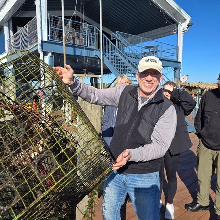 Smiling man lifting a seaweed-covered lobster trap on a sunny marina dock with a blue two-story waterfront building, stairs, and people in the background under a clear blue sky
