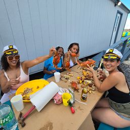 Four friends in captain hats enjoying a dockside summer crab boil at a beachside shack, holding steamed crabs over lemon-sprinkled shells and beer on a picnic table