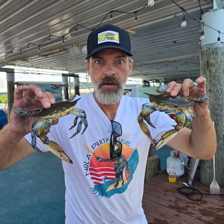Person on a wooden pier holding two live blue crabs with a surprised expression, wearing a cap and New Orleans–themed T-shirt at a waterfront seafood dock