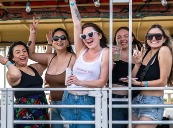 Five friends smiling and cheering on an open-air party trolley during a daytime summer bachelorette celebration, bride-to-be wearing a 'Bride to Be' tank and heart-shaped sunglasses, holding drinks.