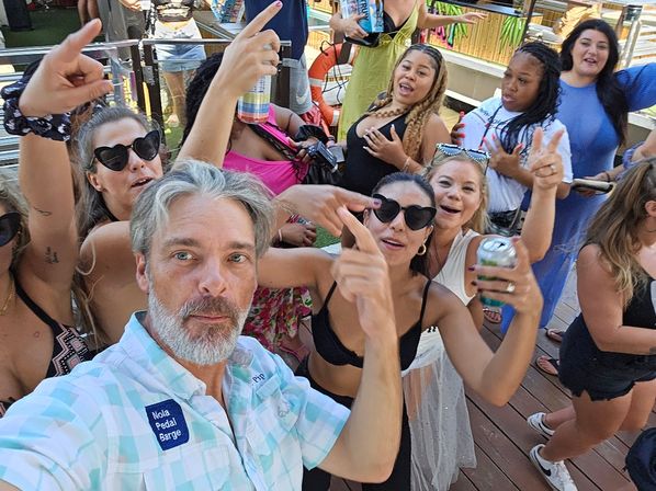Man taking a selfie with a lively group at a summer boat-deck party — people in sunglasses and swimwear holding drinks and dancing on a wooden deck.