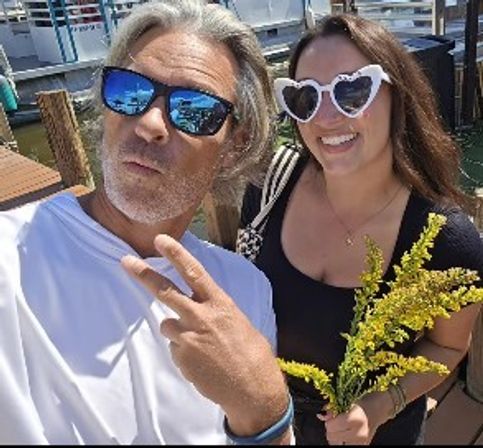 Sunny marina dock selfie: man in mirrored sunglasses flashing a peace sign and woman in heart-shaped shades smiling while holding yellow wildflowers.