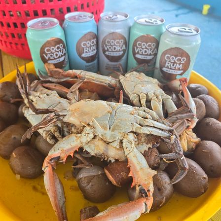 Yellow platter of steamed crabs and baby potatoes with canned coconut cocktails in the background — tropical outdoor seafood boil