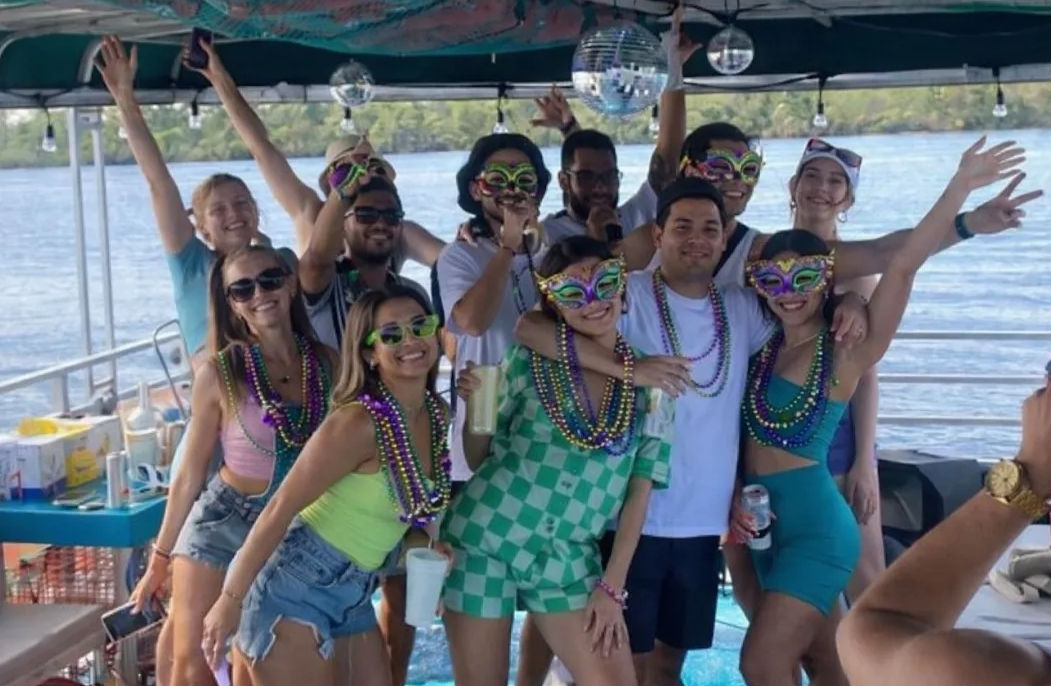 Group of friends on a festive boat party, wearing colorful Mardi Gras masks and layers of bead necklaces, smiling and holding drinks under a decorated canopy on a sunny river cruise.