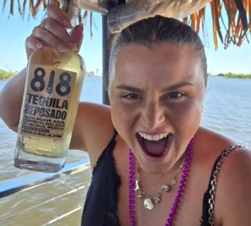 Cheerful woman on a sunny lakeside tiki-style boat wearing purple beads and holding up a bottle of reposado tequila