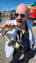 Person in sunglasses and white jacket playfully holding several live blue crabs with claws extended, mouth open in a mock bite at a sunny outdoor seafood festival or waterfront market.