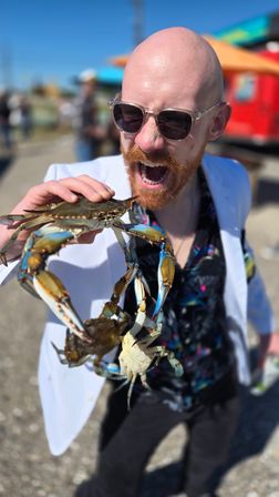 Person in sunglasses and white jacket playfully holding several live blue crabs with claws extended, mouth open in a mock bite at a sunny outdoor seafood festival or waterfront market.