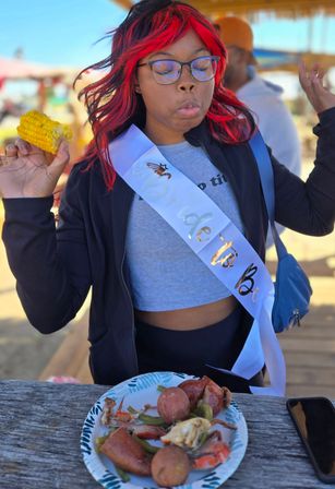 Person with red-streaked hair wearing a "Bride to Be" sash making a playful face while holding a corn on the cob at a beachside picnic table, paper plate of seafood boil with crab, potatoes and sausage in front.