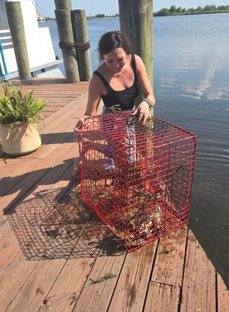Smiling woman on a wooden waterfront dock lifting a red wire crab pot full of shellfish and seaweed, calm bay in the background