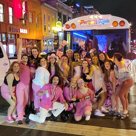 Group of people dressed in pink posing in front of a colorful Party Bus on a lively Nashville nightlife street at night with neon bars and crowds in the background.
