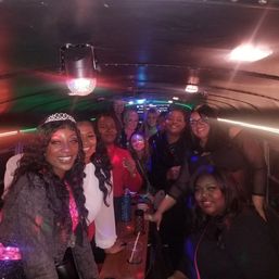 Group of women smiling on a party bus with colorful disco lights, drinks, and a tiara-wearing celebrant enjoying a night out.