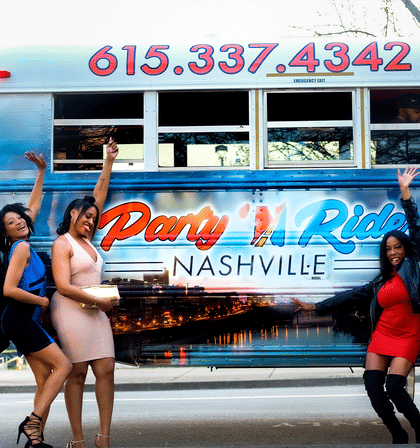 Three women in party dresses cheer and pose beside a colorful Nashville party bus decorated with skyline graphics and bright lettering at a city curb.