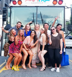 Group of friends striking playful poses and peace signs in front of a Nashville party bus on a downtown street, wearing colorful summer outfits and heels