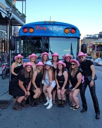 Group of friends wearing pink cowgirl hats and boots posing in front of a blue party bus on a lively downtown Nashville street