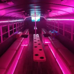 Vibrant pink neon-lit party bus interior with long bench seating, central wooden cup-holder rack, vertical dance pole and a TV screen at the rear