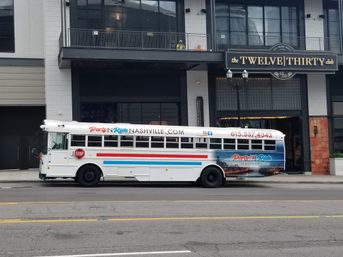 White party bus with red and blue stripes parked on a downtown Nashville street in front of a multi-story entertainment venue with balcony and street lamps