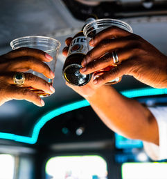 Two hands clinking plastic cups and a beer bottle overhead inside a neon-lit vehicle interior, amber beer pouring in a lively celebratory toast