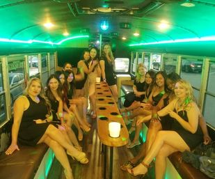 Group of women in black dresses smiling inside a green-lit party bus interior with bench seating, center table of cup holders and a dance pole — girls' night out or bachelorette celebration.