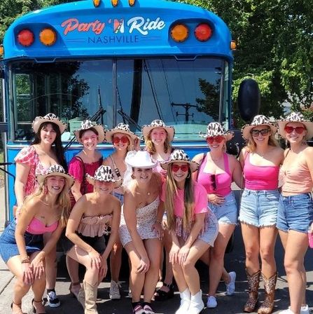 Group of women in pink outfits and cowgirl hats posing and smiling in front of a bright blue party bus on a sunny Nashville street