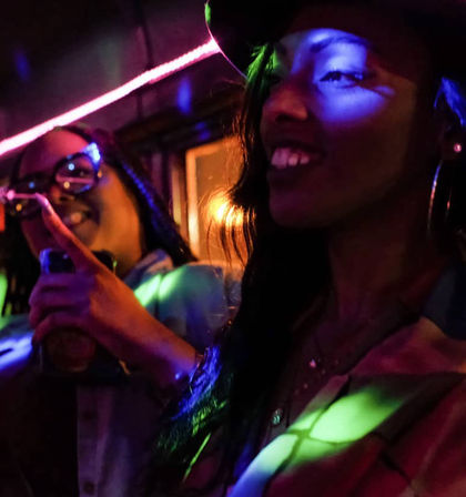 Two friends smiling in a neon-lit lounge, colorful blue and green LED lights casting patterns on their faces while one holds a drink — lively urban nightlife scene.