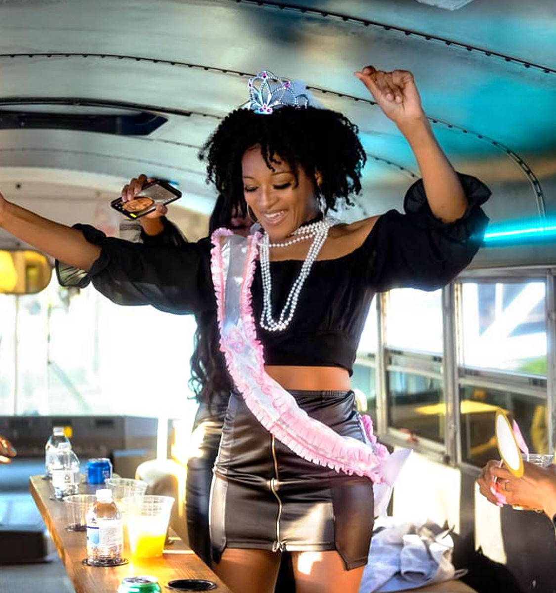 Bachelorette-style celebration on a party bus: person wearing a tiara, pink sash and pearls dancing in a black crop top and leather skirt with drinks on a side table.
