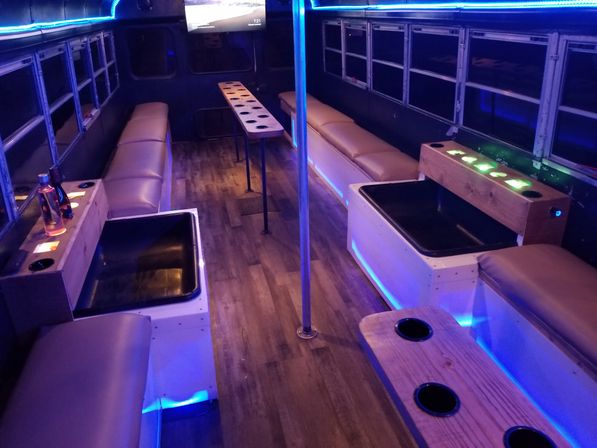 Party bus interior with blue LED accent lighting, leather bench seating, wood-look floor, multiple cup holders, tub footwells, center pole and wall-mounted TV