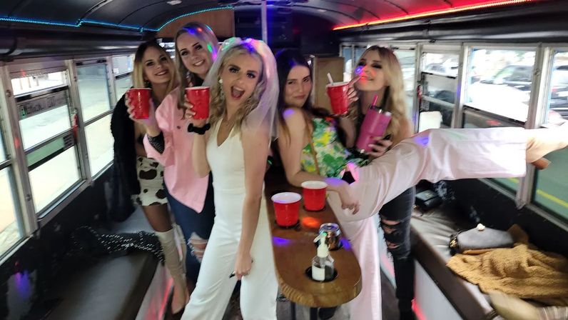 Bachelorette party on a lit party bus — friends holding red cups, bride-to-be in a white dress and veil, colorful LED lights and drinks on a wooden center table.