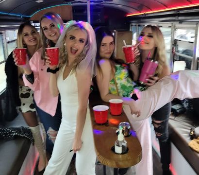 Bachelorette party on a lit party bus — friends holding red cups, bride-to-be in a white dress and veil, colorful LED lights and drinks on a wooden center table.