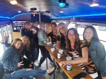 Group of women friends partying inside a neon-blue lit party bus, gathered around a wooden bar table with cans, cups and smiling faces.