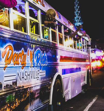 Nighttime Nashville party bus with colorful neon lights and skyline wrap, passengers leaning from windows waving and enjoying a lively street scene