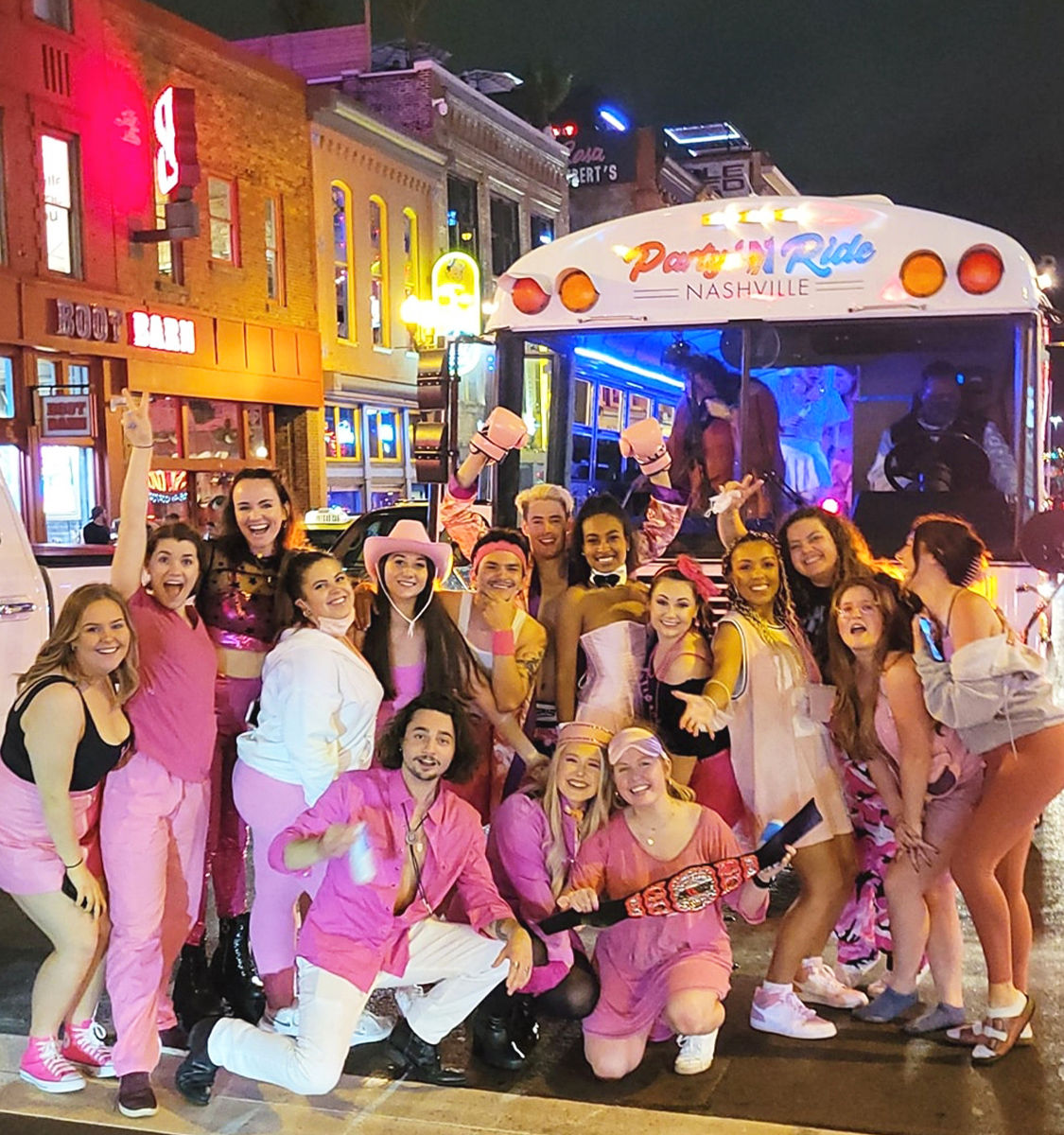 Energetic group in pink outfits cheering and posing in front of a lit party bus labeled “Nashville” on a vibrant downtown nightlife street with neon signs at night