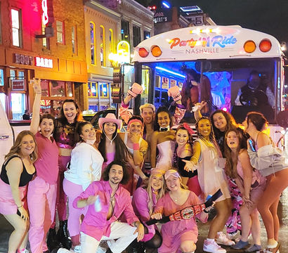 Energetic group in pink outfits cheering and posing in front of a lit party bus labeled “Nashville” on a vibrant downtown nightlife street with neon signs at night
