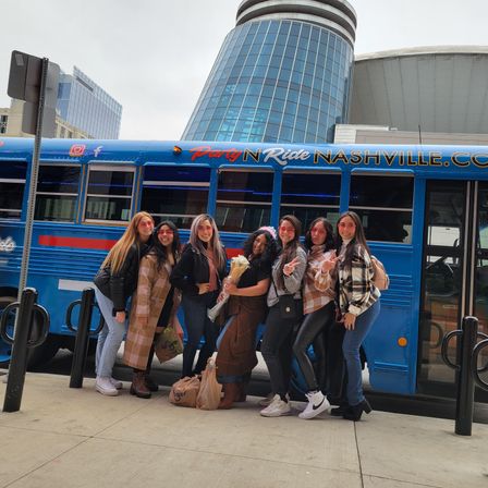 Seven friends smiling and posing in front of a blue party bus in downtown Nashville, one holding a bouquet, wearing coats and sneakers with a modern glass building in the background