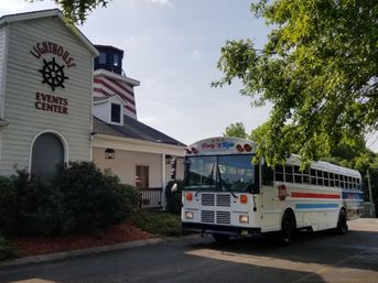 White lighthouse-style event center with a nautical wheel sign beside a parked white party bus with red and blue stripes under leafy trees on a sunny day.