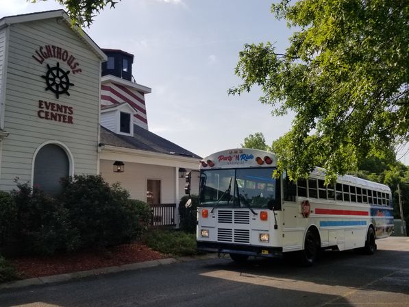 White lighthouse-style event center with a nautical wheel sign beside a parked white party bus with red and blue stripes under leafy trees on a sunny day.