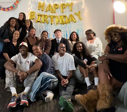 Group of people gathered in a living room for a birthday celebration, smiling on couches and the floor in front of gold “HAPPY BIRTHDAY” balloons with a tinsel backdrop and string lights.