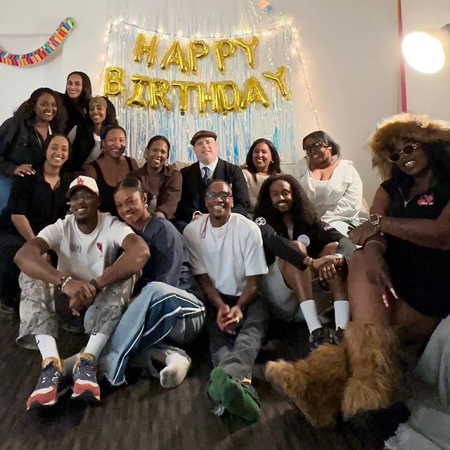 Group of people gathered in a living room for a birthday celebration, smiling on couches and the floor in front of gold “HAPPY BIRTHDAY” balloons with a tinsel backdrop and string lights.