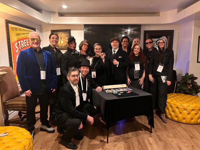 Group photo of thirteen adults in eclectic formal and costume attire gathered in a cozy indoor event lounge around a table with cards, a top hat and feather prop, vintage clock and poster on the wall, and a yellow tufted ottoman.