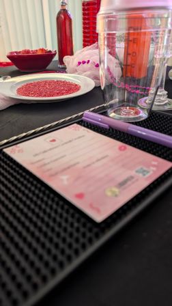 Valentine's-themed table close-up: plate of red glitter sprinkles, red bottles, pink loofah with heart beads, clear cocktail shaker and a purple pen resting on a pink RSVP/menu card.