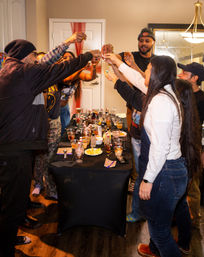 Friends at an indoor house party raising shot glasses for a cheers around a black-covered table filled with cocktails, lemon wedges and drink mixers — lively celebration.