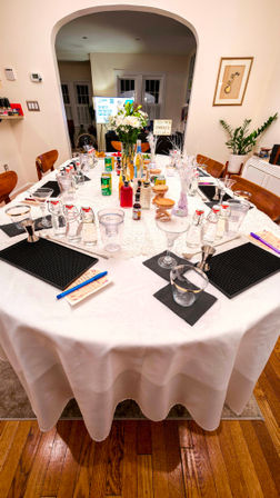 Home dining room table set for a cocktail-making class with martini glasses, mixers, bitters, soda cans, small bottles and jars, notepads and pens, and a white floral centerpiece.