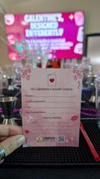 Hand holding a pink "My Galentines Cocktail Creation" recipe card at a cocktail-making station with jiggers, shakers, a red straw and a blurred pink event screen in the background — Galentine’s cocktail workshop vibe