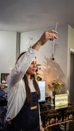 Person in apron holding a long twisted bar spoon aloft in a living room holiday gathering, with a decorated Christmas tree, flowers and a lightbox sign reading 'CONGRATS JOE' on a table.