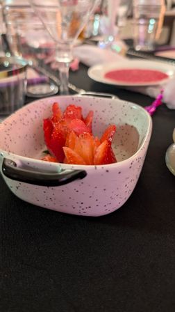 Speckled ceramic bowl holding carved strawberry “flower” garnishes on a black tablecloth, with blurred wine glasses and place settings in the background.