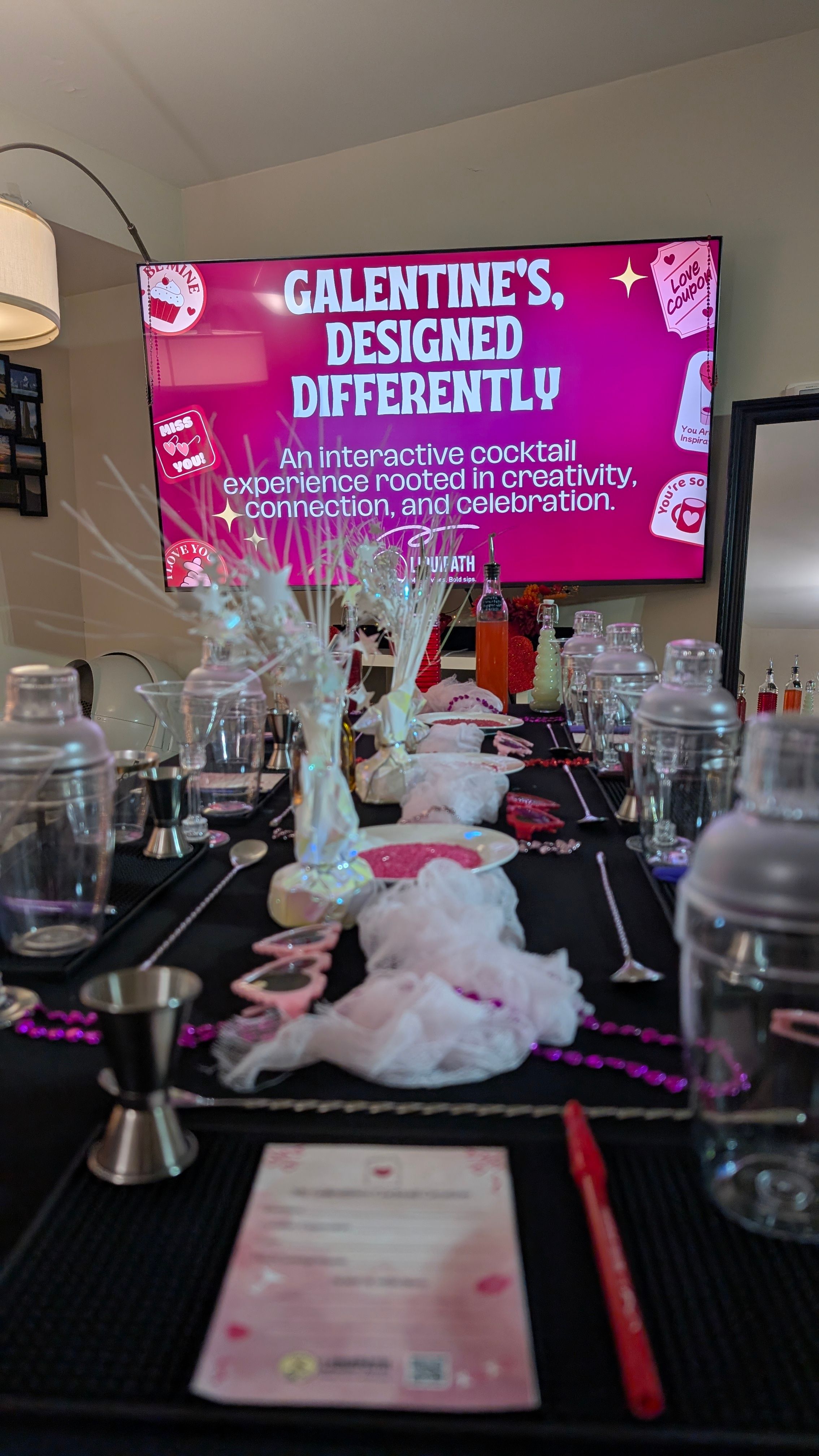 Indoor Galentine's cocktail workshop setup in a living room: pink-themed TV sign, shakers and jiggers, mixing spoons, bowls of pink sugar and sprinkles, syrup bottles, decorative beads and menus.