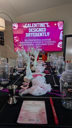 Indoor Galentine's cocktail workshop setup in a living room: pink-themed TV sign, shakers and jiggers, mixing spoons, bowls of pink sugar and sprinkles, syrup bottles, decorative beads and menus.