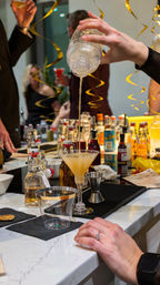 Hand pouring a shaken cocktail into a martini glass at a festive home bar counter with bottles, mixers, and gold streamers.