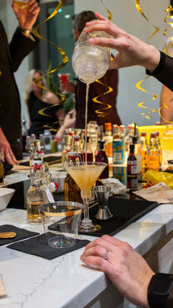 Hand pouring a shaken cocktail into a martini glass at a festive home bar counter with bottles, mixers, and gold streamers.